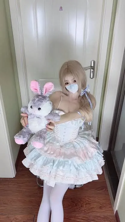 一口白桃七 cosplay海外名称 一口白桃七浅色裙装造型，手持玩偶位于室内背景前