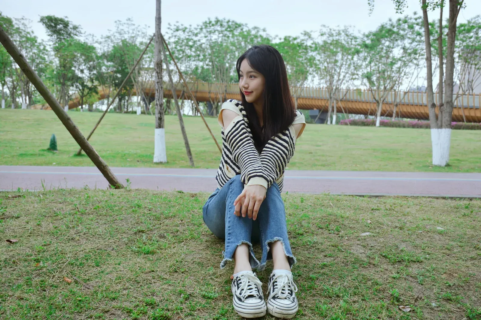 户外公园草坪上身着黑白条纹露肩长袖与牛仔裤的年轻女性坐姿侧拍