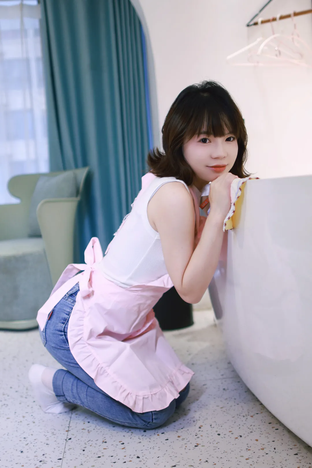 晓晓可可爱跨语言名称识别与Cosplay作品整理 齐刘海短发人物穿白色无袖上衣、粉色围裙和蓝色牛仔裤,跪坐在室内浅色台面旁