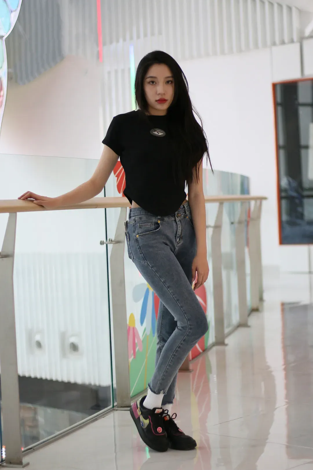 室内商场玻璃护栏旁身穿黑色收腰短袖与修身牛仔裤的长发女性立姿全身照