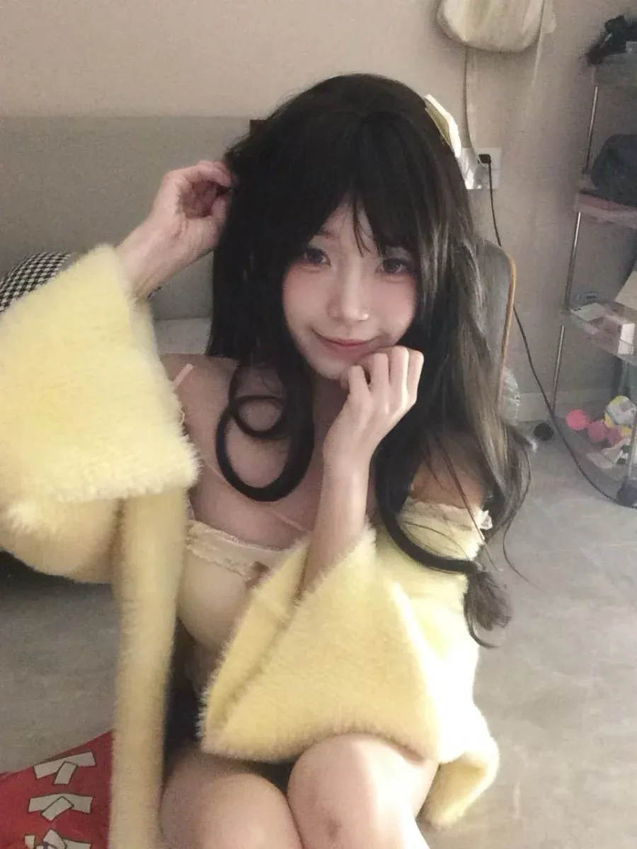 留黑长卷发并身穿明黄色毛绒外套托腮俯视镜头的室内近景