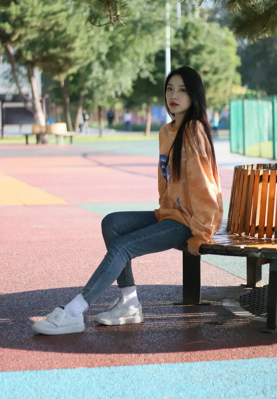 户外彩色地坪休闲区木质弧形长椅上身穿橘黄扎染卫衣与牛仔裤的年轻女性坐姿