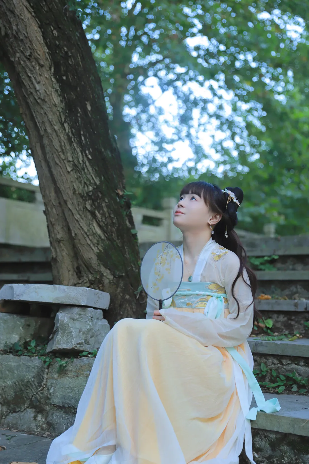 晓晓可可爱跨语言名称识别与Cosplay作品整理 长发盘饰人物穿浅黄白传统服装,手持圆形团扇坐在树旁石阶上并抬头望向上方